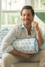 A smiling man with light brown hair and a beard sits on a sofa in a light blue shirt and beige pants, holding the Calico Mini Floral Bolster Pillow adorned with vibrant blue flowers. Sunlight from a window brightens the background.