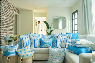 A bright living room with European elegance features a beige couch adorned with Euro Blue Tiles and Spring Blue Stripe Pillow, extra pillows in a floor basket, a brick accent wall, and a person in white walking by in the background.