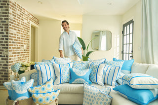 In a bright living room, a person stands next to a white sofa adorned with Yellow Calico Scallops Pillows. A brick accent wall, flowers in a vase, and a large mirror enhance the relaxed and cheerful ambiance.