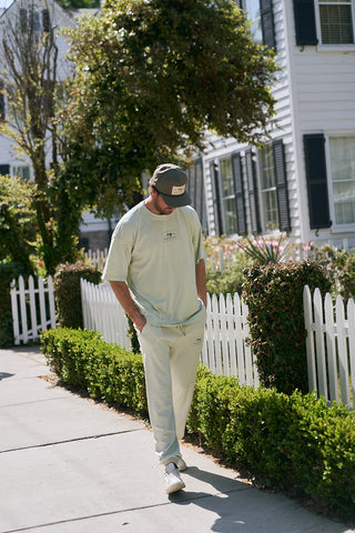 A man wearing The Patch Logo Tee in an oversized boxy fit walks by a white picket fence and garden in a residential neighborhood, paired with matching pants, sneakers, and a dark cap.