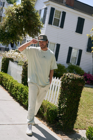 A man in relaxed fit, light-colored heavyweight fleece Patch Logo Joggers and a cap walks on a sidewalk by a white picket fence, with trimmed bushes and a white house in the background.