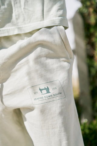 Close-up of The Patch Logo Joggers, relaxed fit light-colored pants with a pocket and a rectangular Sewing Down South patch logo featuring a sewing machine, made from soft cotton fleece and worn outdoors.