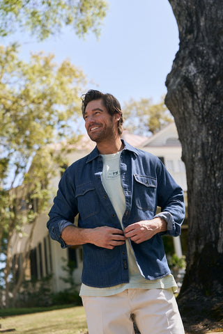 A man stands outside near leafy trees and a sunlit house, smiling as he buttons an oversized blue shirt over The Patch Logo Tee in light green.
