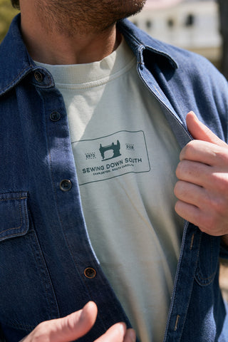A person wears a denim button-up shirt over The Patch Logo Tee, a light-hued heavyweight jersey T-shirt featuring a sewing machine graphic and "Sewing Down South, Charleston, South Carolina" text.