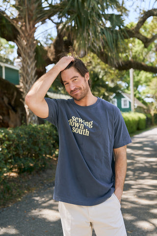 A man stands outside on a sunny day, smiling and touching his hair. He wears The SDS Logo Tee by Sewing Down South—a navy blue retro unisex t-shirt in a relaxed fit, made from soft ring-spun cotton—with trees and houses behind him.