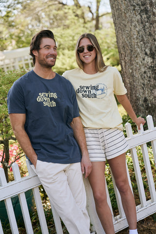 A man and woman smile by a white picket fence outdoors, both wearing The SDS Logo Tee—the man in navy with white pants, the woman in light yellow with striped shorts and sunglasses. Trees and greenery fill the background.