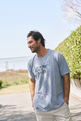 A man with dark hair and facial hair stands outdoors in sunlight, smiling and wearing The SDS Logo Tee—a light blue, unisex relaxed fit shirt made from retro washed ring-spun cotton—with green bushes and water behind him.
