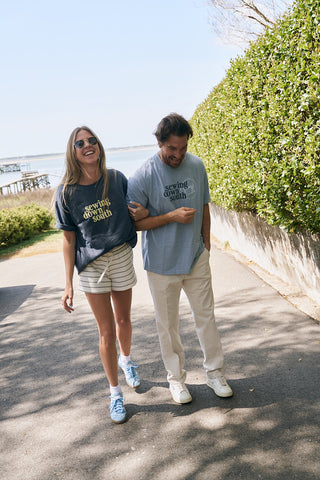 A smiling woman and man walk arm-in-arm outdoors on a sunny day, both wearing The SDS Logo Tee in a retro, relaxed fit. She pairs hers with shorts and sunglasses; he wears light pants. Green bushes line the path.