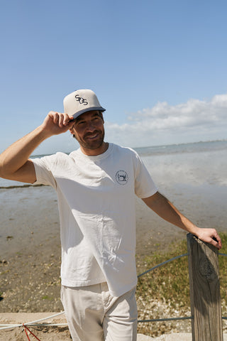A man in a white t-shirt and light pants smiles by the water, holding the brim of The Needle Logo Embroidered Hat. The blue sky, clouds, shoreline, and water are visible in the background.