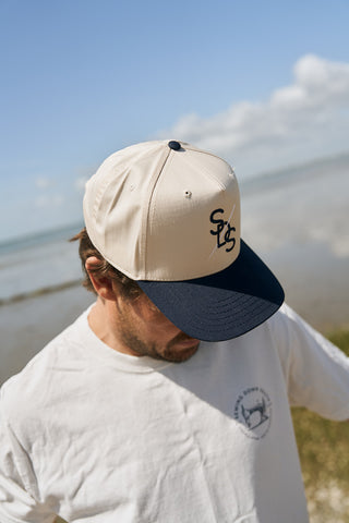 A man stands outdoors by the water, looking down and wearing The Needle Logo Embroidered Hat with a stylized SC emblem, along with a white T-shirt featuring a small round chest logo.