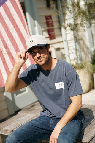 A man in a blue T-shirt and jeans sits on an outdoor bench, tipping The Silicone Patch Hat in Navy. An American flag and plants can be seen in the background.