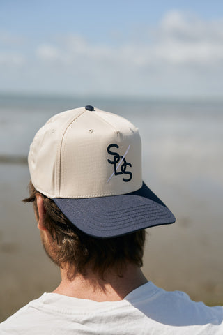 A person with brown hair, seen from behind, wears The Needle Logo Embroidered Hat in beige and navy. The SOLS and diagonal line logo are visible. The background features a blurred seascape with clouds and calm water.