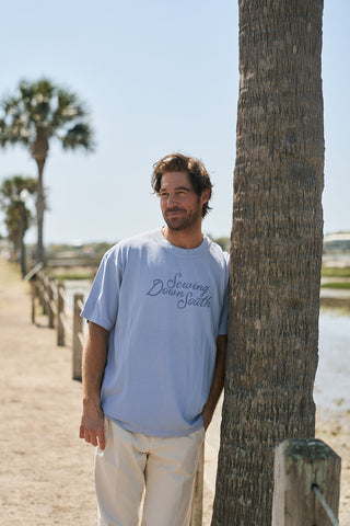 A man in a light blue oversized boxy fit The Rainbow Row Script Tee and light pants leans against a palm tree on a sunny, sandy path with more palms and a wooden fence beside a body of water.