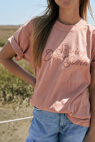 A woman stands outdoors in a field wearing The Rainbow Row Script Tee, an oversized pink boxy-fit shirt with Sewing Down South written on it, and light blue denim shorts. Her left hand rests on her hip.