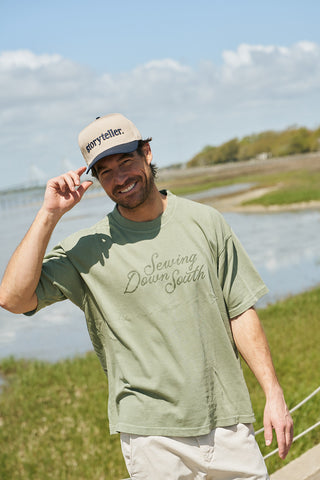 A smiling man by the water wears a khaki cap and The Rainbow Row Script Tee—a loose, heavyweight sage-green jersey with an oversized boxy fit—while holding the cap’s brim on a sunny day.