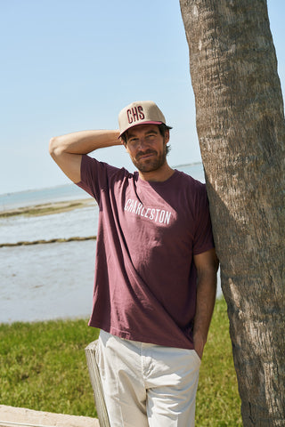 A man in a CHS 3D Embroidered Hat, Maroon, a maroon Charleston t-shirt, and light pants stands by a palm tree on grassy shore with the ocean and blue sky behind him.