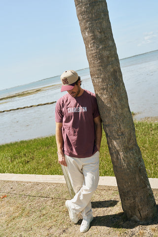 A man in a mauve Charleston t-shirt, beige pants, and a maroon CHS 3D Embroidered Hat leans against a palm tree near the water, looking down with his hands in his pockets on a sunny day.