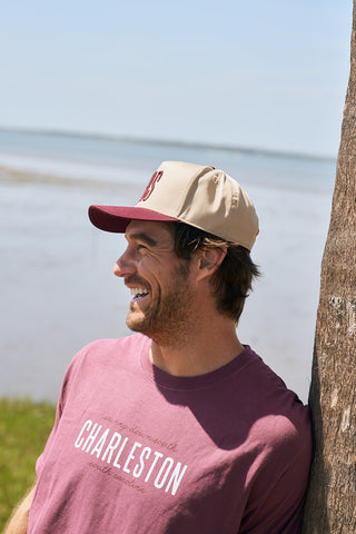A smiling man in a maroon CHS 3D Embroidered Hat leans against a tree by the water, wearing a maroon CHARLESTON shirt. The scene features a peaceful shoreline and blue sky in the background.