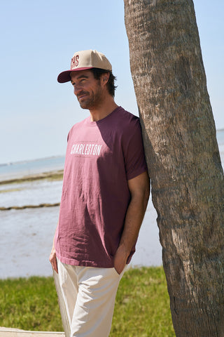 A man in a relaxed fit, burgundy The Charleston Tee, beige pants, and a beige cap smiles while leaning against a palm tree by the water on a sunny day, with marshland and blue sky in the background.