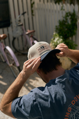 A person wearing a blue shirt adjusts The Silicone Patch Hat, Navy while standing outdoors near a pink bicycle and a white fence, with sunlight casting shadows on the scene.