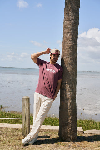 A man in The Charleston Tee and light pants leans against a palm tree by a calm shoreline, touching the brim of his hat and smiling on a sunny day.