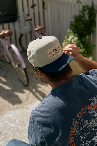 A person wearing The Silicone Patch Hat, Navy, and a blue t-shirt with a sun graphic sits outdoors near a white fence and two pink bicycles, adjusting their hat while facing away from the camera.