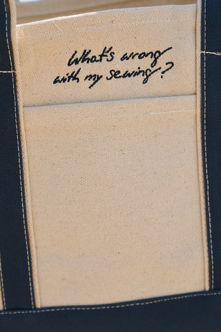 A close-up of The WWWMS Embroidered Tote, featuring Craig's Handwriting with "What's wrong with my sewing?" on a beige pocket and dark navy edges.