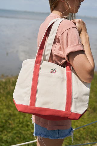 A person in a pink shirt and denim shorts stands by the water, carrying the Heirloom Tomato Embroidered Tote Bag—a large cotton canvas bag with red straps and a small embroidered cherry design.
