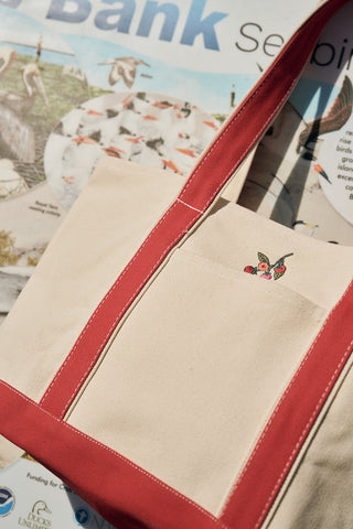 The Heirloom Tomato Embroidered Tote Bag—beige cotton canvas with red straps and a small tomato design—rests on an illustrated magazine or newspaper featuring birds and the word "Bank" in bold blue letters.