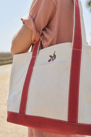 Wearing a pink shirt, a person carries the Heirloom Tomato Embroidered Tote Bag, a cream canvas tote with red straps and embroidered cherry design, set against a sunny backdrop that reflects the Garden Prep Capsule Collection’s outdoor inspiration.