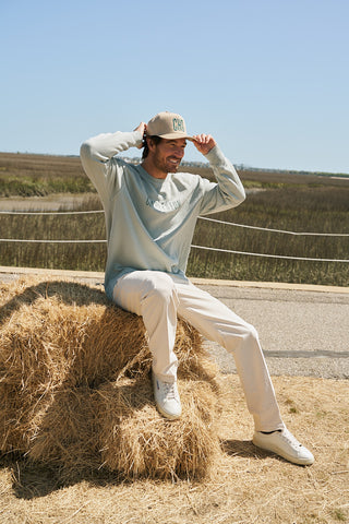 A man in a light blue shirt, beige pants, white sneakers, and a CHS 3D Embroidered Hat in Teal sits on hay bales outdoors, smiling on a sunny day with fields and a road in the background.