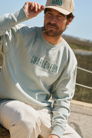 A man in The Charleston Crewneck Sweatshirt, light blue classic fit fleece, and a beige cap sits outside on a sunny day, holding straw in his mouth and lifting his hat.