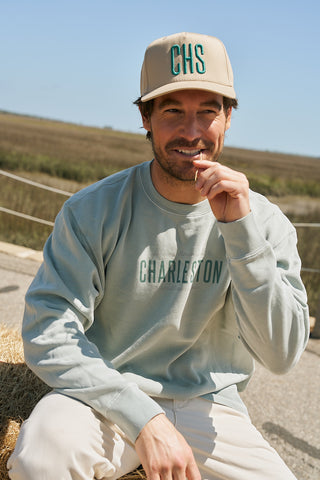 A man in a teal CHS 3D Embroidered Hat and light blue Charleston sweatshirt smiles outdoors on a sunny day, straw in his mouth, with marshland behind him—perfect scenery for enjoying some local honey.