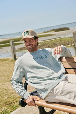 A man wearing a teal CHS 3D Embroidered Hat and a light blue Charleston sweatshirt smiles on a bench by the water, with grass, a wooden fence, and blue sky in the background.