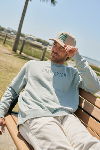 A man in a teal CHS 3D Embroidered Hat and a CHARLESTON sweatshirt relaxes on an outdoor wooden bench surrounded by palm trees and sand, enjoying the day after beekeeping and tasting local honey.