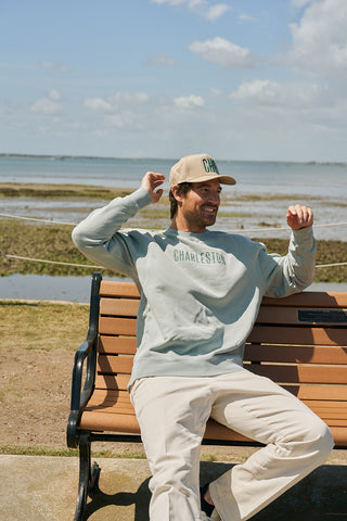 A man in a light green The Charleston Crewneck Sweatshirt and a beige Charleston cap sits smiling on a park bench by the water, with grass, sand, and blue sky in the background.