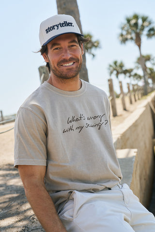A bearded man smiles outdoors under palm trees in a sunny sky, wearing The WWWMS Tee—a relaxed fit, retro unisex ring-spun cotton shirt featuring "What's wrong with my sewing?"—and a white Storyteller cap.