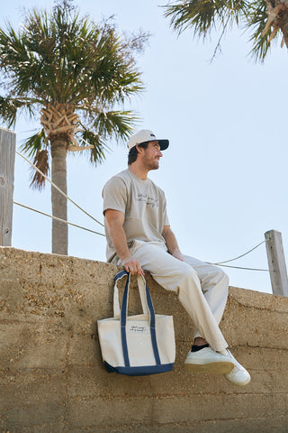 A man in a beige t-shirt, light pants, and white sneakers sits on a stone wall holding The WWWMS Embroidered Tote with Craig's Handwriting; palm trees and clear blue sky fill the background.