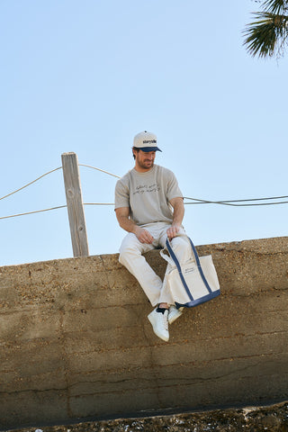 A man in a beige T-shirt, white pants, and white sneakers sits on a concrete wall holding The WWWMS Embroidered Tote. He wears a beige cap and looks down, with a clear blue sky and palm tree in the background.