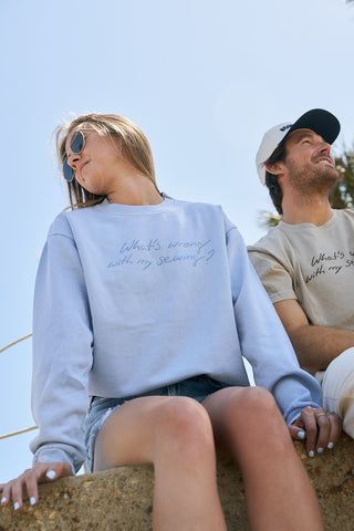 A woman in sunglasses and The WWWMS Crewneck Sweatshirt, a light blue pigment-dyed top with an oversized boxy fit and "What's wrong with my sewing?" text, sits outdoors beside a man in a beige shirt and white cap.