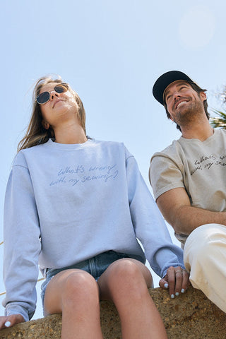 Two people sit outdoors smiling. One wears sunglasses, shorts, and The WWWMS Crewneck Sweatshirt in light blue with "Whats wrong with my sewing?" text; the other wears a cap, beige T-shirt, and light pants.