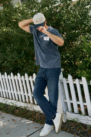 Wearing jeans, a blue T-shirt, and white sneakers, a person stands before a white picket fence with greenery behind, adjusting The Silicone Patch Hat in Navy.