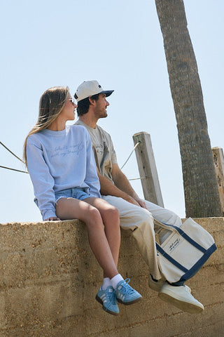 A woman wears The WWWMS Crewneck Sweatshirt in light blue with an oversized, boxy fit and shorts, sitting on a low wall beside a man in beige shirt and cream pants. A tote bag lies on the ground near them, with a palm tree in the background.