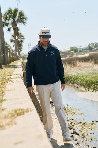 A man wearing The Needle Logo Embroidered Quarter Zip, a storyteller cap, and beige pants walks along a sunny waterside path with palm trees and grass in the background.