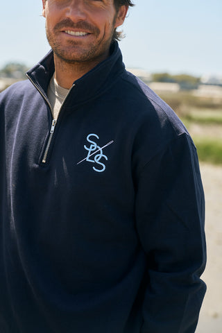 A man stands outdoors, smiling, in The Needle Logo Embroidered Quarter Zip—navy blue cotton-polyester fleece with white SXS initials on the left chest, a durable metal zipper, and standard fit. Blurred greenery and sand fill the background.