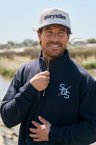 A smiling man wearing The Needle Logo Embroidered Quarter Zip in navy blue stands outdoors near a marsh, partially zipping up his beige "storyteller" cap.