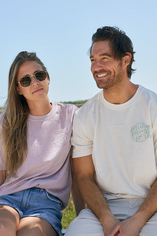 A woman in sunglasses and a pink The Stamp Tee sits next to a smiling man in a white tee outdoors under a clear blue sky. Both look relaxed and casual.