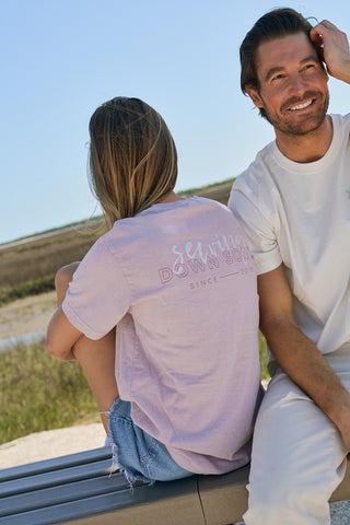 A woman in a relaxed lavender tee and denim shorts sits on a bench with her back to the camera. Beside her, a smiling man enjoys the outdoors in The Stamp Tee, a light-colored retro unisex shirt, near a grassy marsh under clear skies.