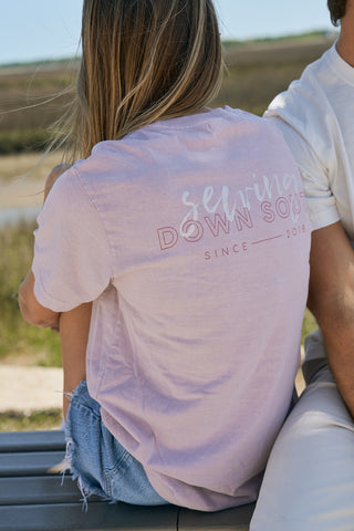 A person with long blonde hair wearing The Stamp Tee—a relaxed fit, light purple Sewing Down South retro unisex t-shirt—and denim shorts sits on a bench outdoors beside another person in a white shirt.