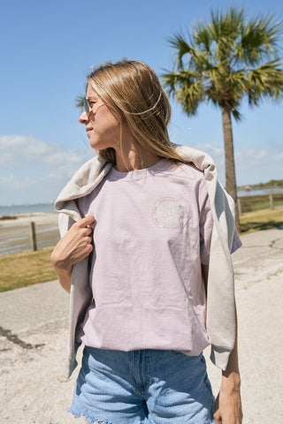A woman in The Stamp Tee—light purple, relaxed fit, retro unisex—paired with denim shorts and sunglasses stands outdoors by a palm tree, a sweatshirt draped over her shoulders on a sunny day.